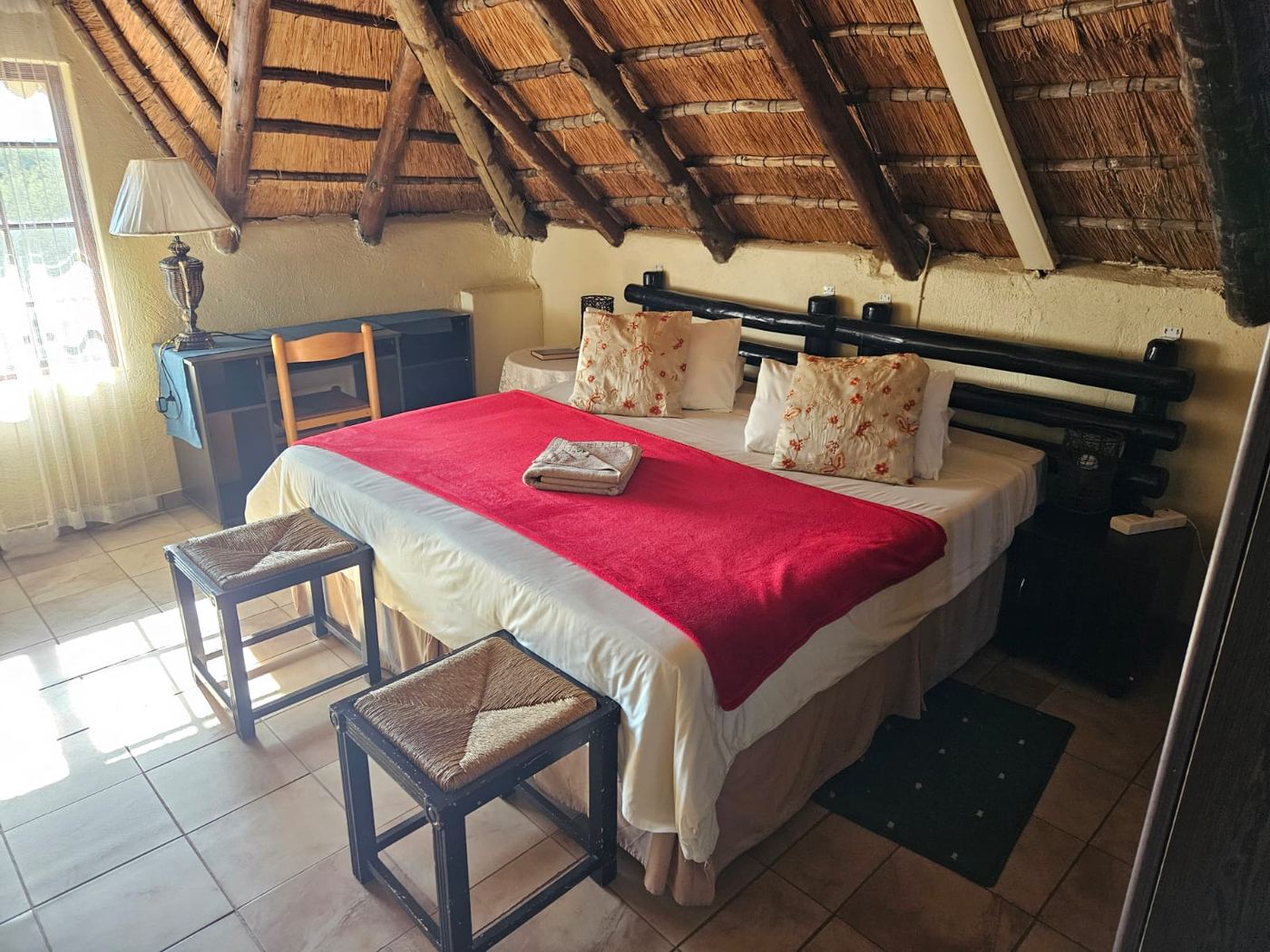 Double room under a thatched roof with exposed poles, red throw and writing desk